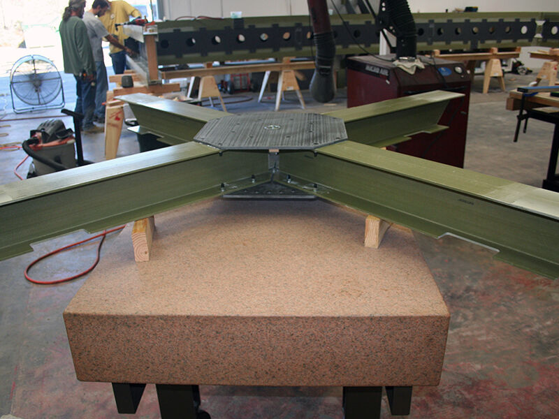 Image shows a cross piece of the test fixture setup on a granite table.  The table is in a manufacturing space with activity in the background, including technicians inspecting part of the long part of the fixture. 